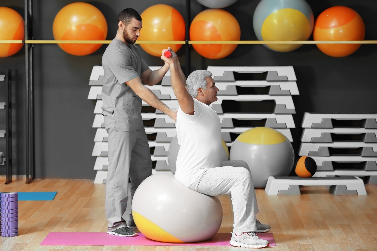 A man in grey scrubs assisting an older man in white clothing who is sitting on a large exercise ball and lifting a small red weight