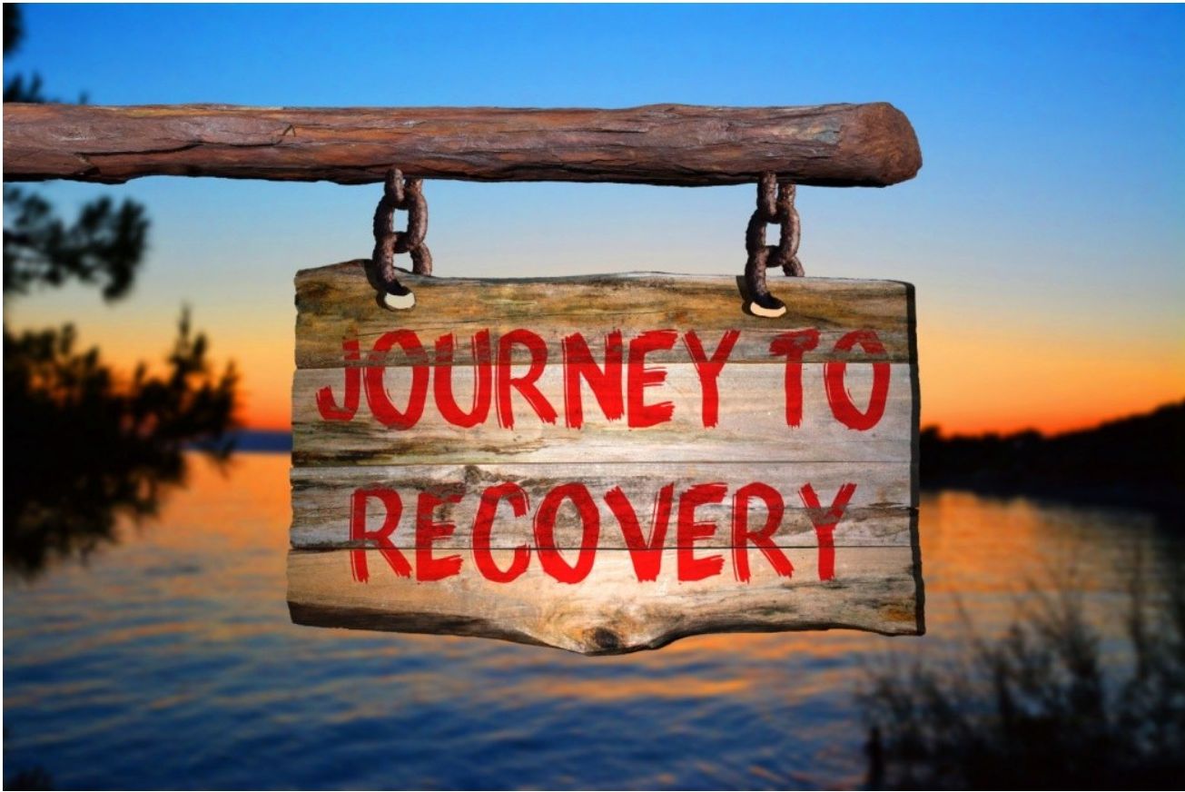 A wooden sign with the words JOURNEY TO RECOVERY painted in red, set against a sunset