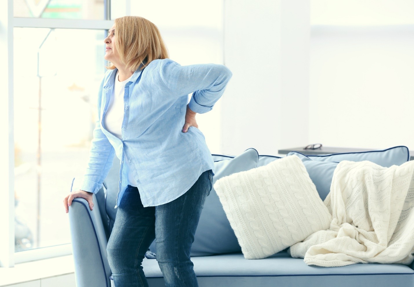 A woman standing by a sofa holding her lower back in pain