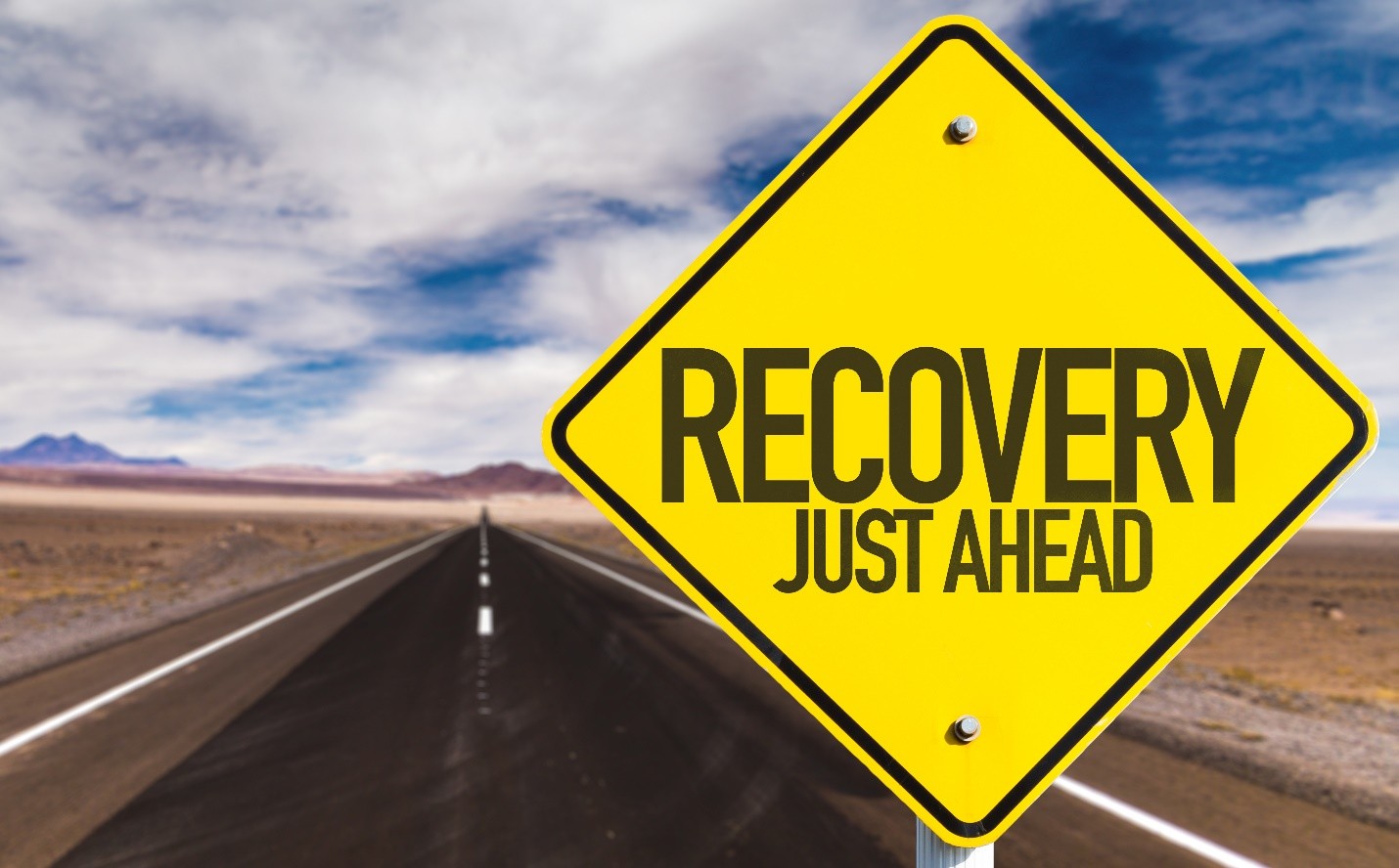 Yellow road sign reading RECOVERY JUST AHEAD on a desert road