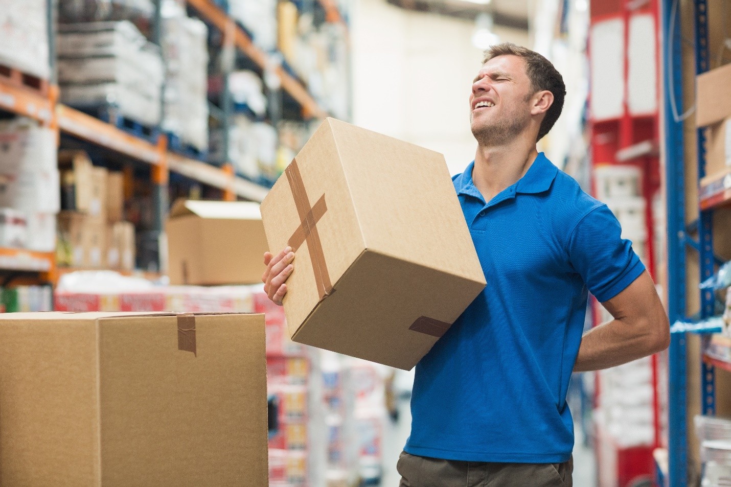 Man in a warehouse holding a cardboard box and clutching his lower back in pain