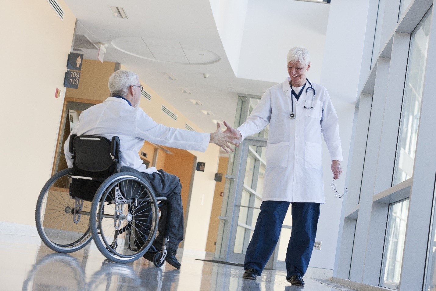 A doctor shaking hands with a patient in a wheelchair in a hospital hallway