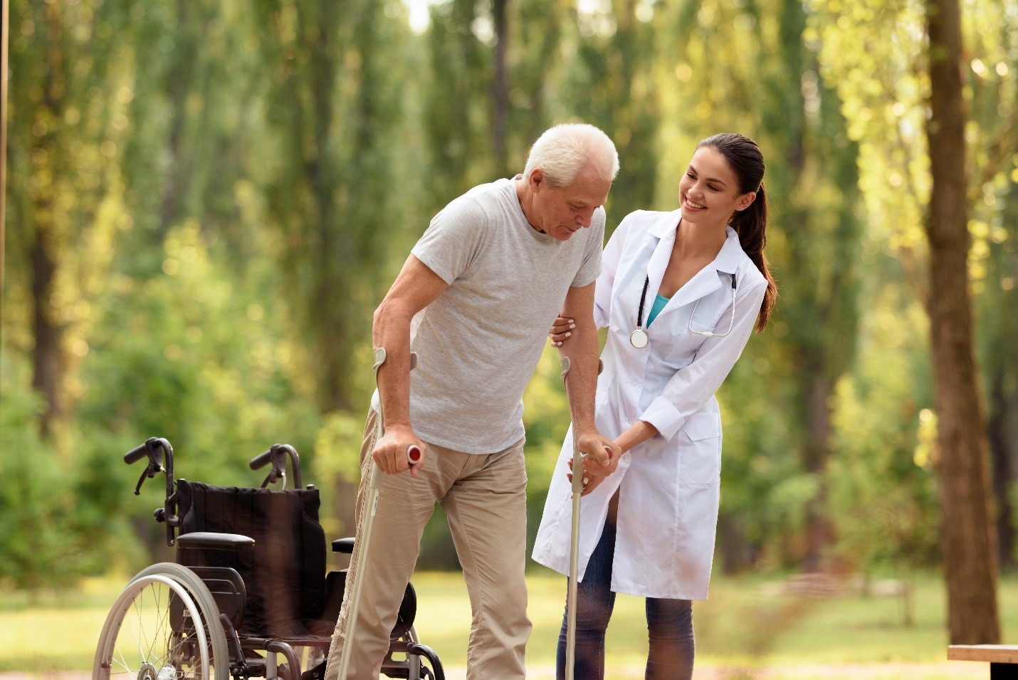 A healthcare professional assisting an elderly man with a cane in a park, with a wheelchair nearby